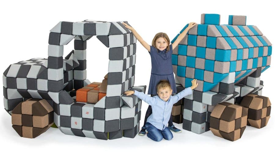 Two children with raised hands standing by a digger and a lorry built from grey and blue JollyHeap blocks