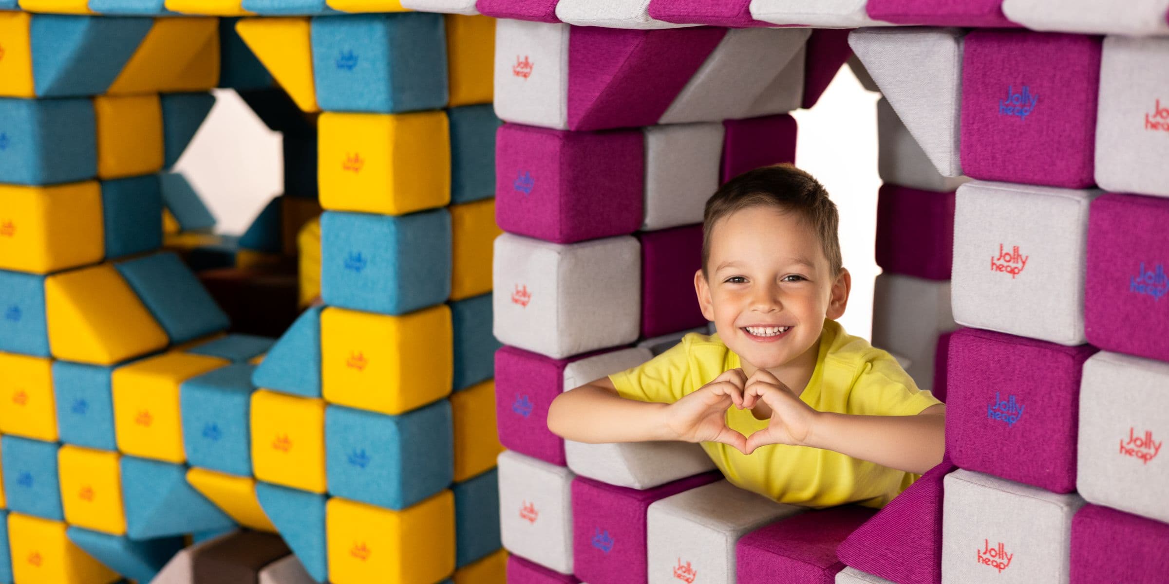 A boy in a yellow t-shirt makes a heart with his hands, looking out of the window of a house built from JollyHeap blocks