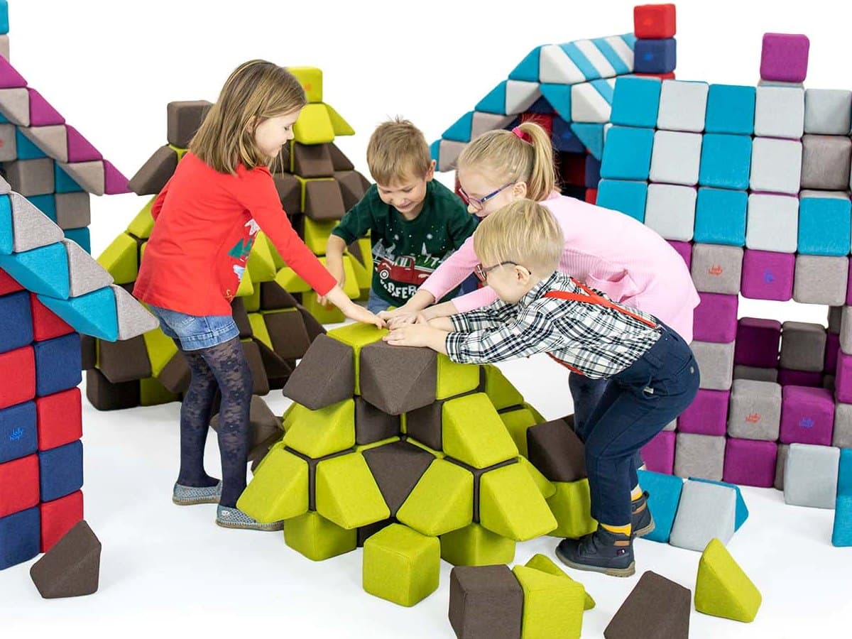 Four children leaning over a shared structure of green and brown JollyHeap blocks, with block-built houses behind them